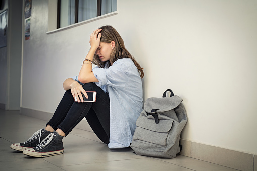 Upset and depressed girl holding smartphone sitting on college campus floor holding head. University sad student suffering from depression sitting on floor at high school. Lonely bullied teen in difficulty with copy space. Middle School therapy blog post, katy Texas, 77494.